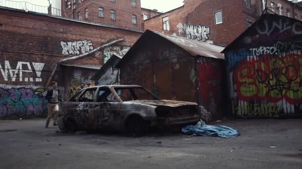Hombre saltando sobre el techo del coche estrellado. Grafitis no identificables en las paredes alrededor — Vídeo de stock