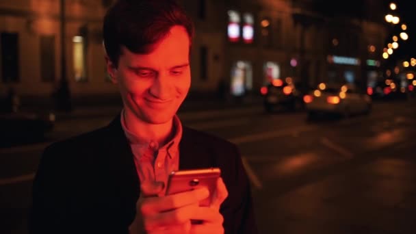 Uomo sorridente che usa lo smartphone di notte in città. Viso illuminato da luce rossa — Video Stock