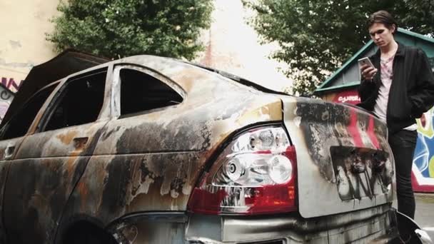 Jeune homme utilisant un smartphone debout près de voiture brûlée écrasée. Mode de vie urbain — Video