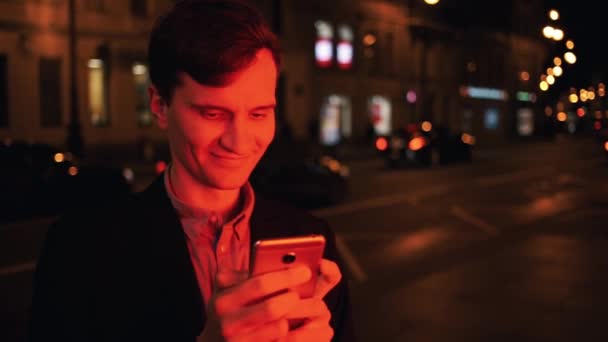 Sourire vêtu d'un jeune homme décontracté utilisant un smartphone la nuit en ville — Video