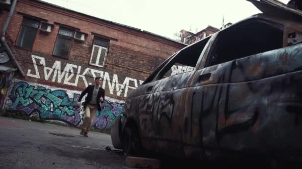 Hombre vestido casual saltando sobre el coche estrellado. Grafitis no identificables en paredes — Vídeo de stock