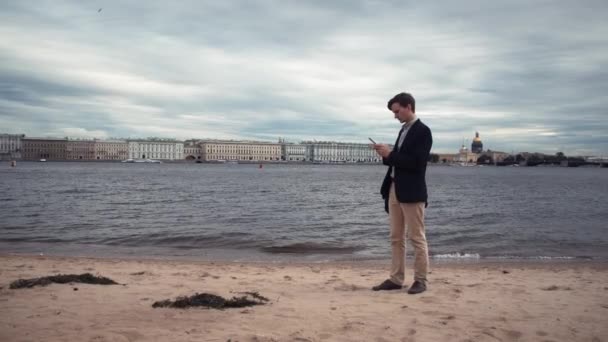 Casual gekleed jongeman met behulp van laptop zittend op de dijk. Buitenshuis werken — Stockvideo