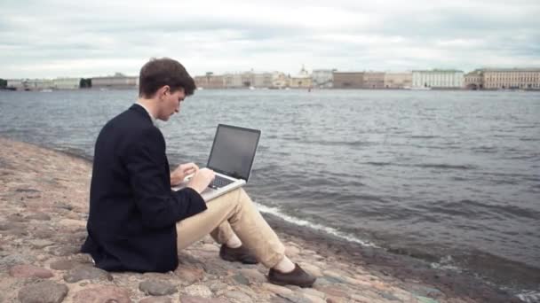 Freelancer met behulp van laptop zittend op een bankje in de buurt van de kust. Zomerdag — Stockvideo