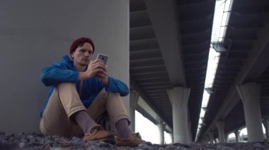 Beyaz delikanlı toprak yolu köprüsü altında oturan smartphone kullanarak