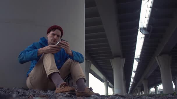 Hombre joven usando un teléfono inteligente sentado en el suelo bajo el puente de carretera . — Vídeo de stock