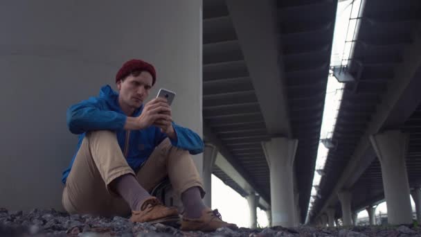 Fiatal természetjáró használ smartphone ülő a földön közúti híd alatt — Stock videók