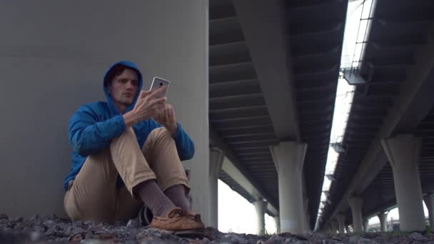 Νεαρός ταξιδιώτης χρησιμοποιώντας smartphone που κάθονται στο έδαφος κάτω από την γέφυρα του δρόμου — Αρχείο Βίντεο