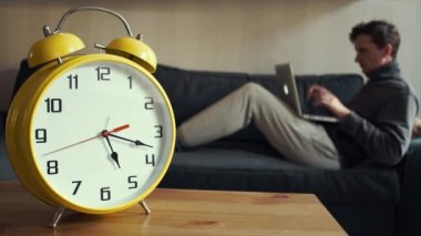 Evde kanepede otururken dizüstü kullanan genç adam. Büyük sarı Saat işliyor