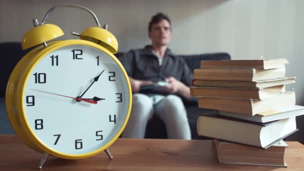 Man met laptop zitting op Bank lezen boek thuis. Gele klok tikt — Stockvideo