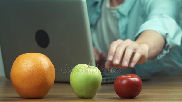 Апельсин, яблоко и помидор на столе перед человеком, который пользуется ноутбуком — стоковое видео