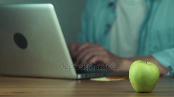 Junger Mann mit einem Laptop, der eine Nachricht tippt. grüner Apfel auf dem Tisch. — Stockvideo