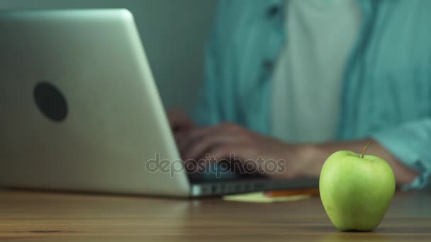 Jovem usando um laptop digitando uma mensagem leva uma maçã verde da mesa — Vídeo de Stock