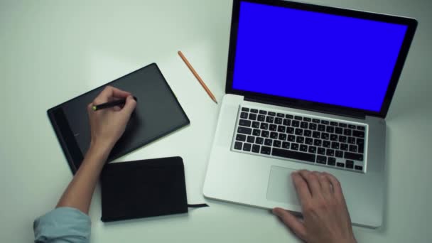 Вид зверху чоловічі руки за допомогою графічної вкладки та ноутбука з зеленим екраном на білому столі — стокове відео