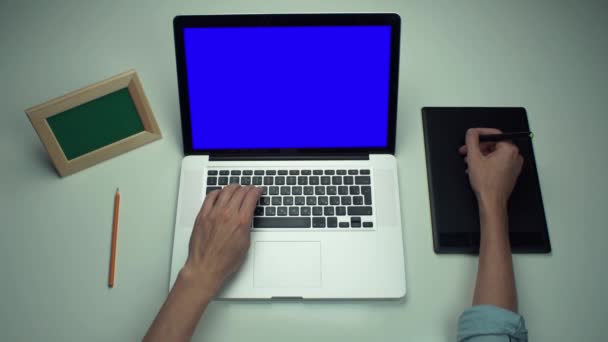 Человек с цифровой графической вкладкой и ноутбук с зеленым экраном на белом столе — стоковое видео