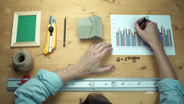 Joven trabajando con gráficos impresos haciendo notas con rotulador en escritorio de madera — Vídeos de Stock