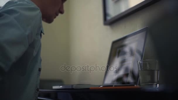 Un joven que usa un portátil en casa. Vaso de agua, bloc de notas en la mesa . — Vídeos de Stock