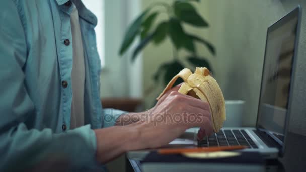 Молодий чоловік їсть банан, використовуючи ноутбук вдома. Чоловік відпрацьовує офіс . — стокове відео