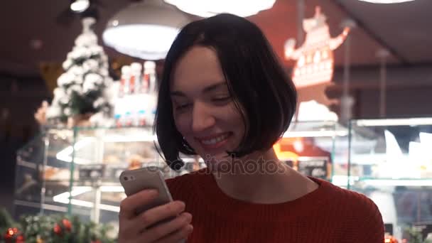 Feliz joven mujer sonriendo mensaje de texto utilizando el teléfono inteligente sentado en la cafetería . — Vídeos de Stock