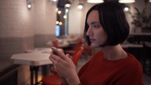 Linda chica joven que usa un mensaje de texto de jersey de punto usando un teléfono inteligente en la cafetería — Vídeos de Stock