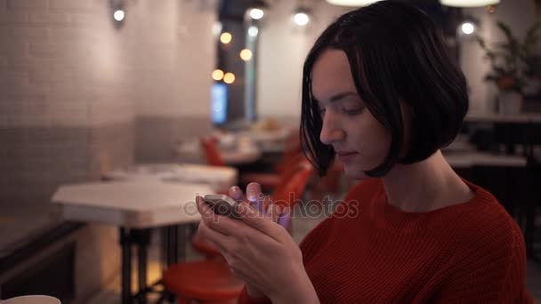 Chica joven caucásica usando teléfono inteligente sentado en la cafetería por la noche — Vídeos de Stock