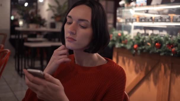 Привлекательная молодая девушка чатится в Интернете с помощью приложения для смартфонов, сидя в пустом кафе — стоковое видео