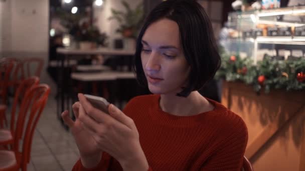 Милая молодая девушка в чате онлайн с помощью приложения для смартфонов, сидящая в кафе вечером — стоковое видео