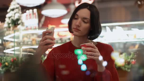Молодая женщина с помощью смартфона делает селфи, сидя в кафе за окном — стоковое видео