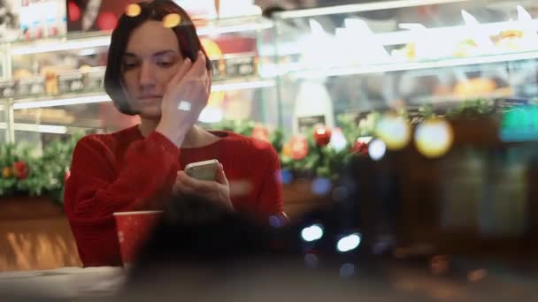 Hermosa chica joven utilizando el teléfono inteligente sentado en la cafetería detrás de la ventana. Tiempo de Navidad — Vídeos de Stock