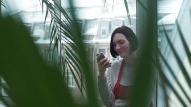 Kırmızı önlük serada smartphone kullanarak gülümseyen genç kadın.