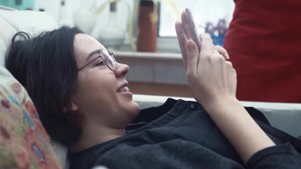 Chica joven sonriendo con el teléfono inteligente tumbado en el sofá en casa . — Vídeos de Stock