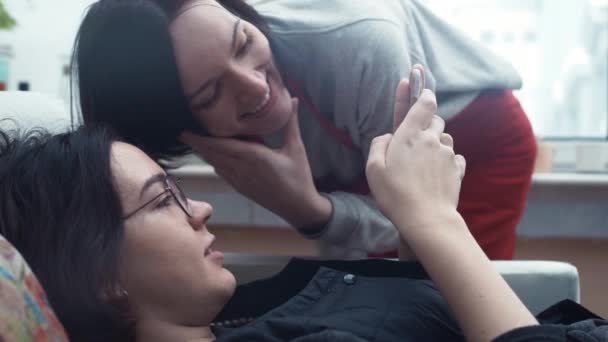 Пара молодых девушек смотрят видео, используя смартфон, лежащий дома на диване — стоковое видео