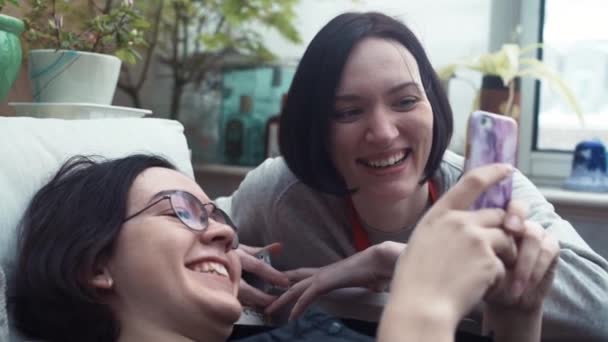 Пара юных подружек смотрят видео, используя смартфон, лежащий на диване — стоковое видео