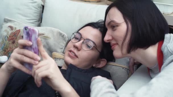 Couple young girls watching video using smartphone lying on couch at home — Stock Video