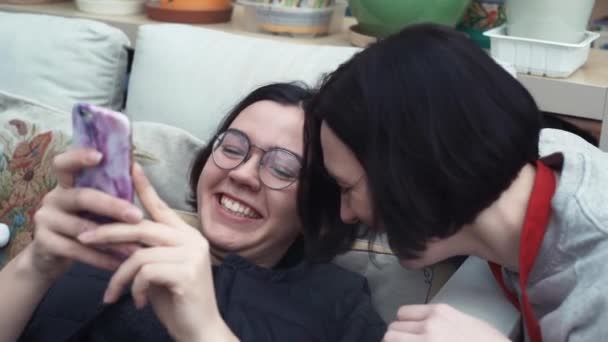 Пара молодих дівчат сміється, використовуючи на смартфоні лежачи на дивані вдома — стокове відео
