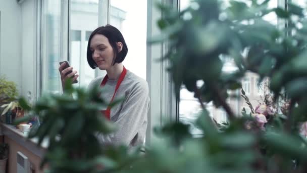 Linda chica en delantal rojo sonriendo con smartphone en invernadero . — Vídeos de Stock