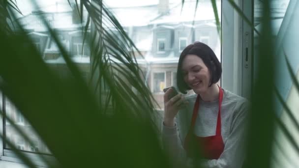 Chica joven en delantal rojo sonriendo con teléfono inteligente en el jardín en casa — Vídeo de stock