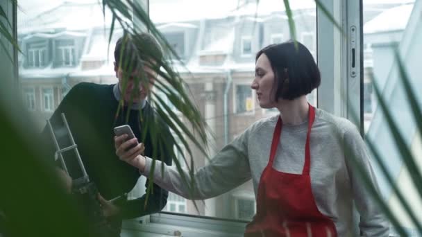Mädchen in roter Schürze lächelt mit Smartphone und zeigt Kerl im Gewächshaus Fotos — Stockvideo