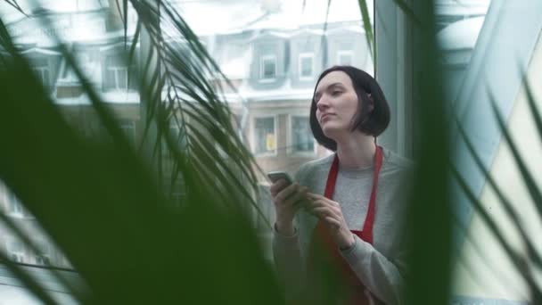 温室効果でスマート フォンを使用して笑みを浮かべて赤いエプロンの若い女性. — ストック動画