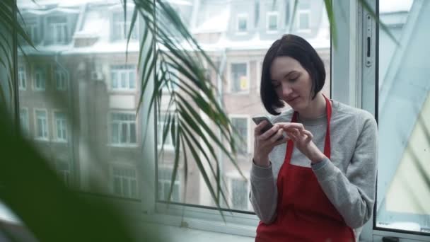 红色围裙的年轻妇女园丁微笑使用智能手机在温室 — 图库视频影像