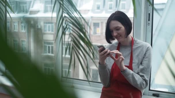 Jonge vrouw tuinman in rode schort glimlachend gebruikend smartphone in kas — Stockvideo