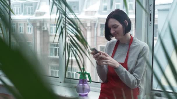Ung söt flicka i rött förkläde leende med hjälp av smartphone i växthus — Stockvideo