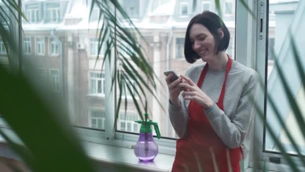 Chica joven usando smartphone en invernadero contra el telón de fondo de la ventana panorámica — Vídeos de Stock