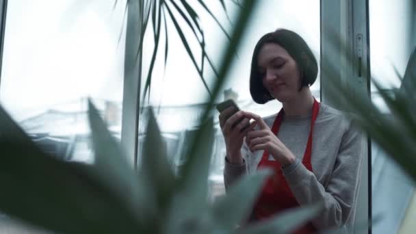 Schattig meisje in de rode schort glimlachend gebruikend smartphone in kas — Stockvideo