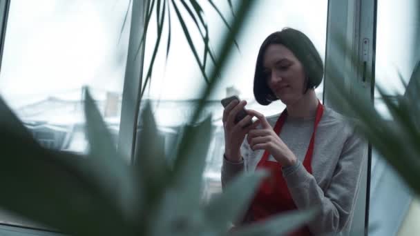 Молодая девушка в красном фартуке улыбается, используя смартфон в саду — стоковое видео