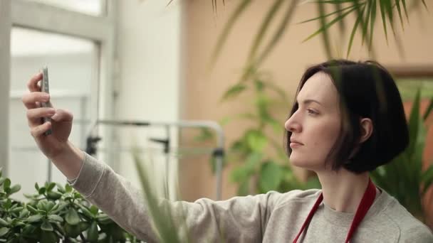Joven linda chica en delantal rojo tomando selfie usando smartphone en invernadero — Vídeos de Stock