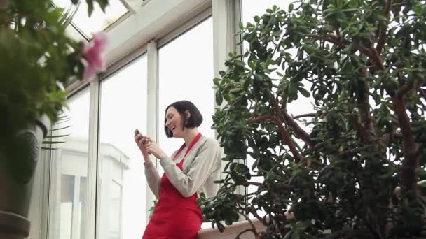 Attraktiv kvinna i rött förkläde skrattar med smartphone i växthus på balkong — Stockvideo