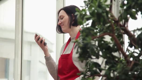 Retrato de mujer atractiva en delantal rojo usando smartphone en invernadero — Vídeo de stock