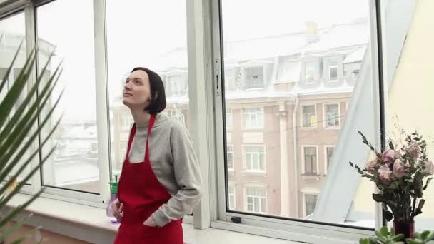 Young woman gardener standing in greenhouse on balcony in front of big window. — Stock Video