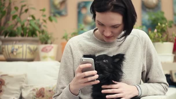 Giovane ragazza utilizzando smartphone seduto con simpatico cane chihuahua nero in soggiorno — Video Stock