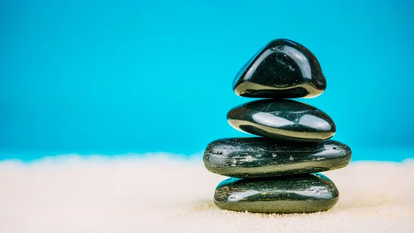 Growing piled up of four black pebbles on bright sand with blue background — Stock Photo, Image